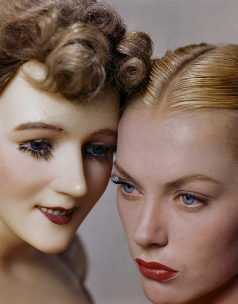 Model cheek to cheek with a beauty-shop mannequin, both seen in ultra close-up. The mannequin is sporting an older curly hairstyle, while the model's hair is styled in the new slicked straight style.
