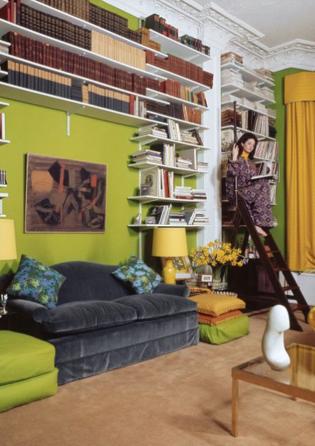 Socialite and hostess Pamela Colin (aka Lady Harlech & Mrs. Ormsby-Gore) seated on an old English library ladder in the drawing room of her London flat/apartment. The room is decorated in bright green and yellow, with a Jean Arp marble on the glass table in the left foreground; shelves of books over a grey sofa which has a painting over it. She wears a patterned jumpsuit over a yellow turtleneck. *** Local Caption *** Pamela Colin;