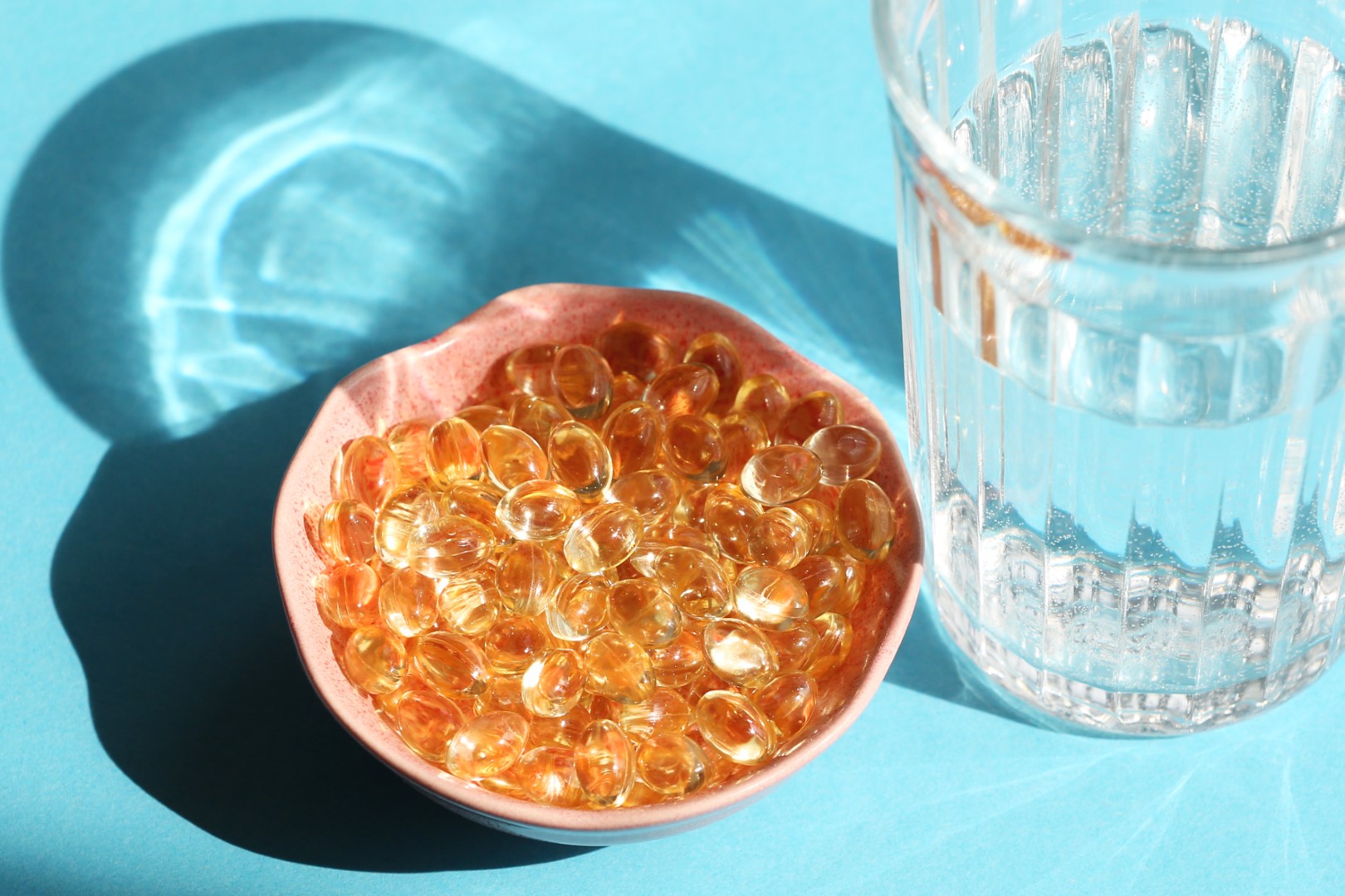 Nutritional Supplements as Vitamin D Capsules in Small Pink Ceramic Plate Next The Transparent Glass Of Water On Blue Background. Healthcare and Alternative Medicine, Immune System and Recovering, Sustainable Lifestyle. Still Life, Natural Lighting, Copy Space