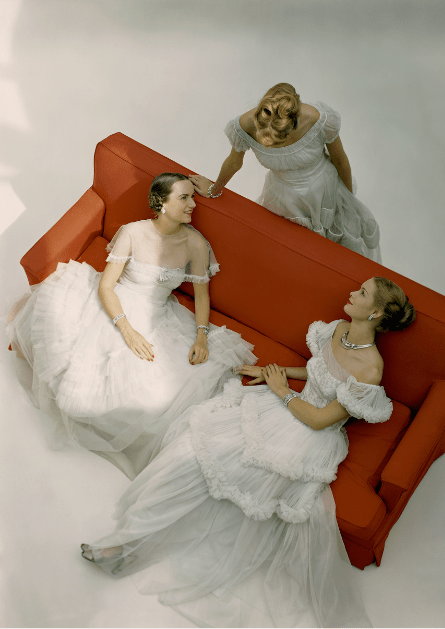 Three models with a red sofa, all wearing white ballgowns for the winter season. The model sitting on the sofa, to the left, wears a full-skirted, ruffle-tiered ball gown of white rayon net, with a small filmy fringed shoulder-cape; styled with silver and rhinestone jewelry. The model standing behind the sofa wears a bright-white rayon net dress with dropped puff-sleeves and pointed bodice, and a skirt of wide corded bands of shirring; styled with a white bracelet. The model sitting on the sofa, to the right, wears an off-the-shoulder ball gown in spangled white rayon net, with ruffles on the sleeves and skirt; styled with a thick silver necklace and bracelet. The gowns are available at Bergdorf Goodman, I. Magnin, and Himelhoch's. All models wear face powder by Schiaparelli, in "Radiance." *** Local Caption ***