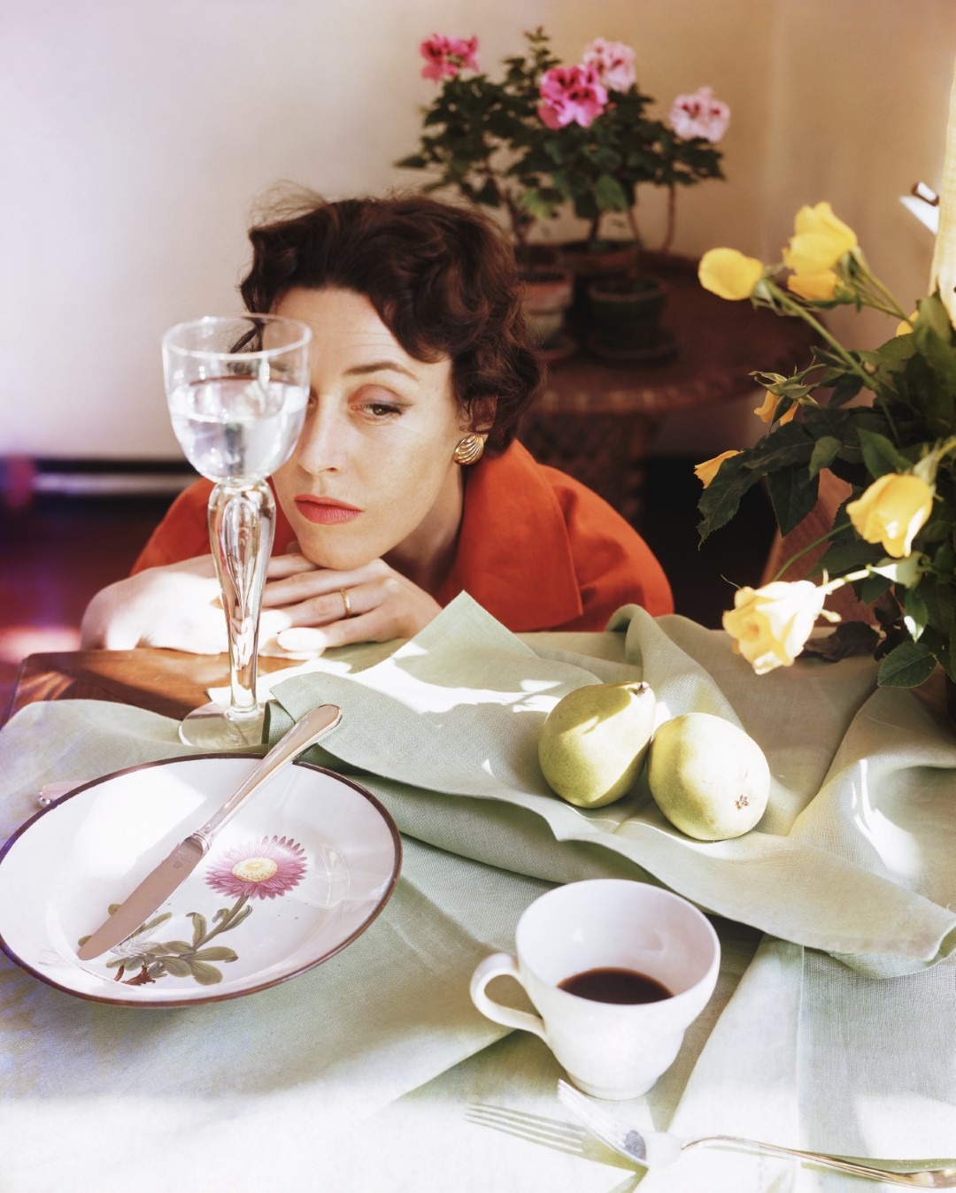 Woman resting her chin on her hands which are resting on a table that is set with an empty plate, a glass of water, a cup of coffee and two pears on a napkin, c.1955