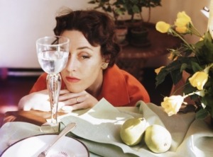 Woman resting her chin on her hands which are resting on a table that is set with an empty plate, a glass of water, a cup of coffee and two pears on a napkin, c.1955
