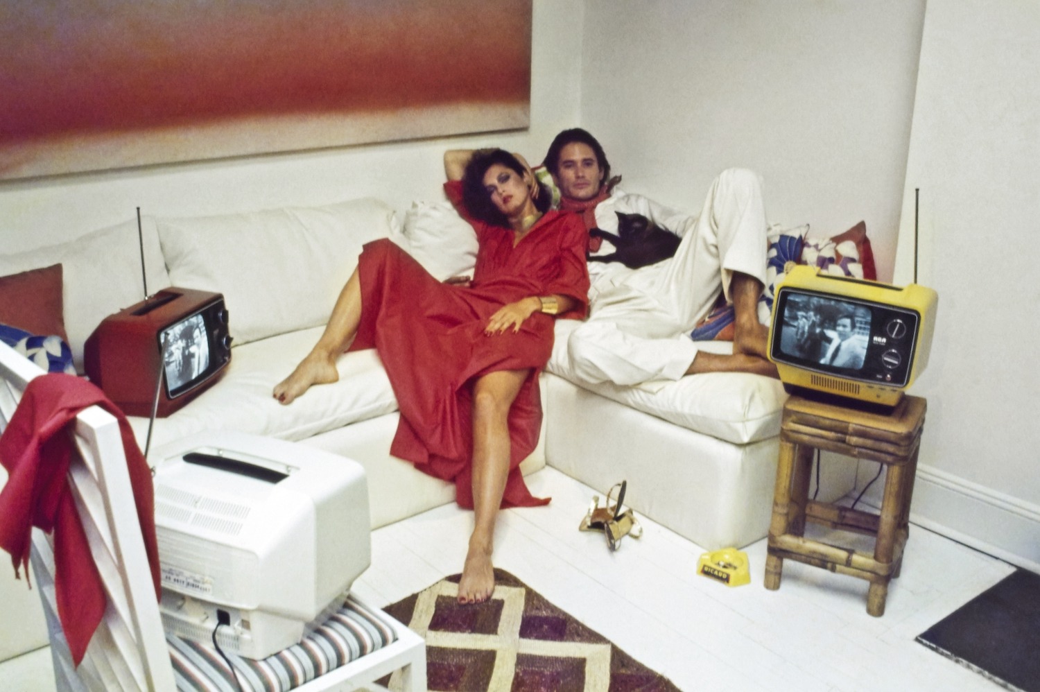Model Dayle Haddon and male model reclining on a white sofa, watching TV (RCA's Sportable 9" screen in three different colors ). She is wearing Fernando Sanchez's red cotton caftan-style dress with high side slit, gathered at the waist, of Verron fabric.  Styled with Berrin Designs earrings, Dionne Cole necklace, cuff by M&J Savitt, and shoes from Margaret Jerrold for Shoe Biz (on the floor next to her feet). Male model wears a white shirt and pants with a red polka-dot scarf. Hair by Harry King. Makeup by Sandra of Xavier Coiffures. Photographed in the New York City apartment of Mark Sephton and Arthur Scott.  *** Local Caption *** Dayle Haddon;