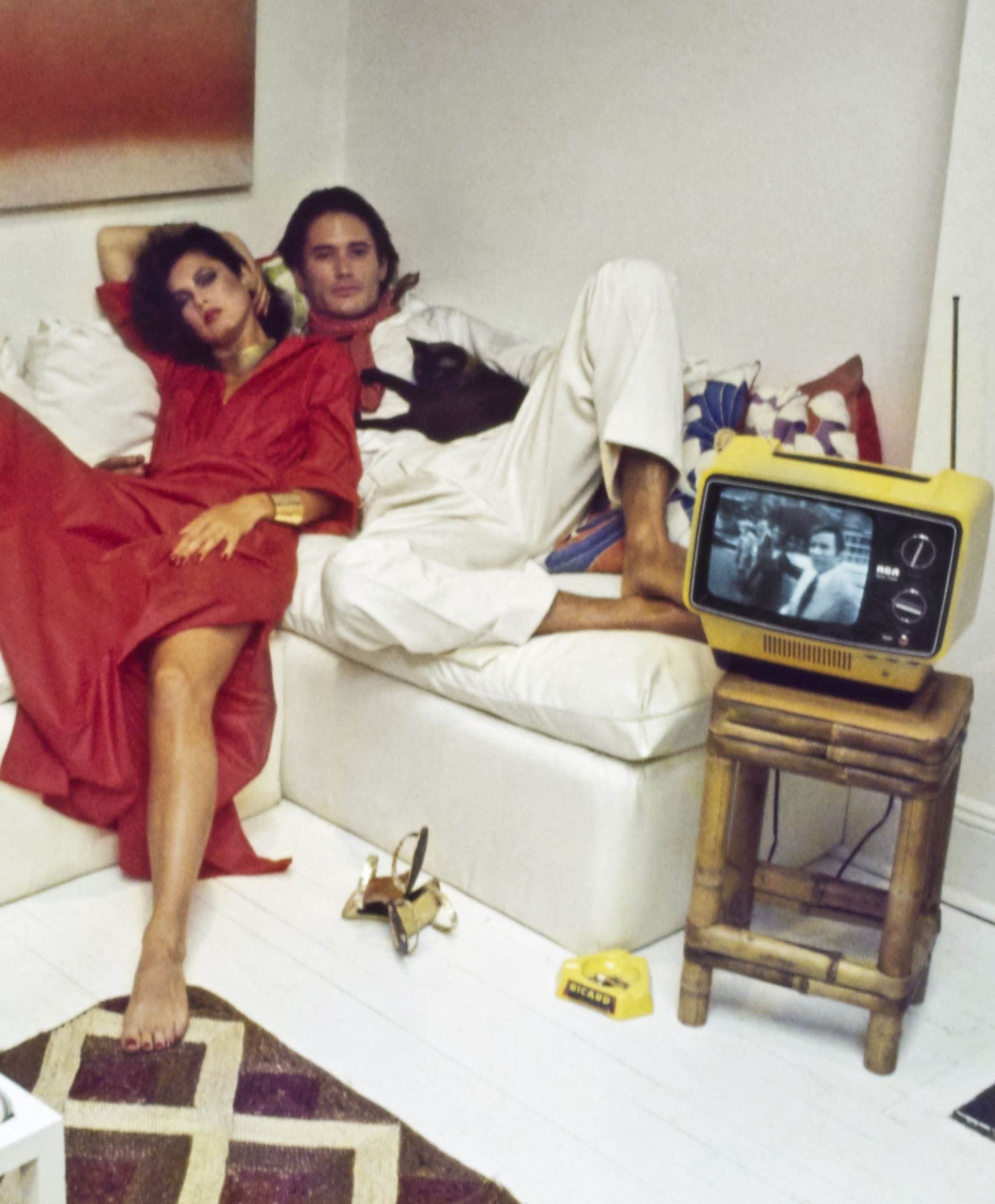 Model Dayle Haddon and male model reclining on a white sofa, watching TV (RCA's Sportable 9" screen in three different colors ). She is wearing Fernando Sanchez's red cotton caftan-style dress with high side slit, gathered at the waist, of Verron fabric. Styled with Berrin Designs earrings, Dionne Cole necklace, cuff by M&J Savitt, and shoes from Margaret Jerrold for Shoe Biz (on the floor next to her feet). Male model wears a white shirt and pants with a red polka-dot scarf. Hair by Harry King. Makeup by Sandra of Xavier Coiffures. Photographed in the New York City apartment of Mark Sephton and Arthur Scott. *** Local Caption *** Dayle Haddon;