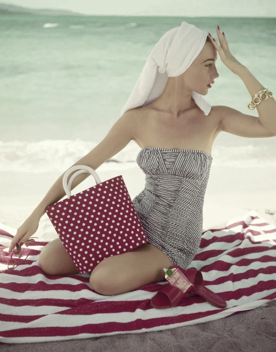 Model seated on a striped beach towel on a beach, wearing a strapless skirted cotton swimsuit, built in panels of gray and white striping (diagonal on the flanks, horizontal down the front of the torso, and vertical across the chest), by Jantzen, with bracelets of linked golt rings by Monet and a white-on-red polka-dotted bag with white handles by MM. Vogue May 01, 1954 Fashion Feature