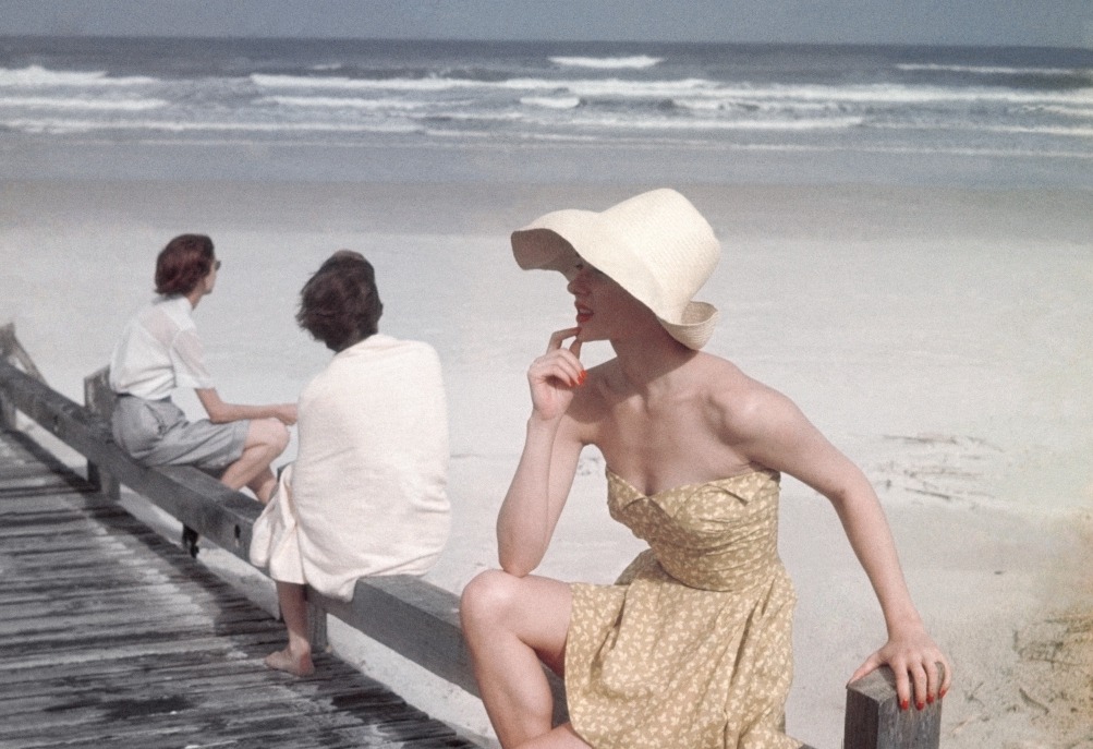 A model seated on a boardwalk at the beach, wearing a yellow petal-top bathing suit, with boning at the waist, by Carolyn Schnurer, of Everfast cotton. Styled with yellow high-laced leather sandals with toe thongs, by Capezio, of Colonial patent leather; a curved-brim natural palm bonnet from the Pan American Shop; and a yellow silk and straw handbag by Koret.