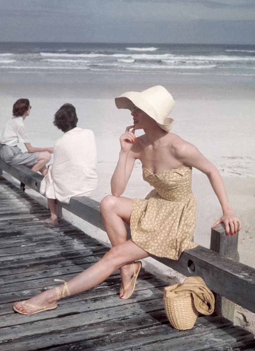 A model seated on a boardwalk at the beach, wearing a yellow petal-top bathing suit, with boning at the waist, by Carolyn Schnurer, of Everfast cotton. Styled with yellow high-laced leather sandals with toe thongs, by Capezio, of Colonial patent leather; a curved-brim natural palm bonnet from the Pan American Shop; and a yellow silk and straw handbag by Koret.