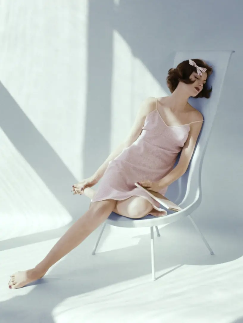 Model Dorothea McGowan sitting in a white molded Fiberglass plastic chair, with a high-back, by Laverne. She wears a pink-and-white gingham print slip with white cording and a scalloped hem, by Asha, of Stoffel fabric of Dacron and cotton. She also has a small white printed bow in her hair.  Dorothea McGowan;Vogue June 01, 1960 Fashion Feature