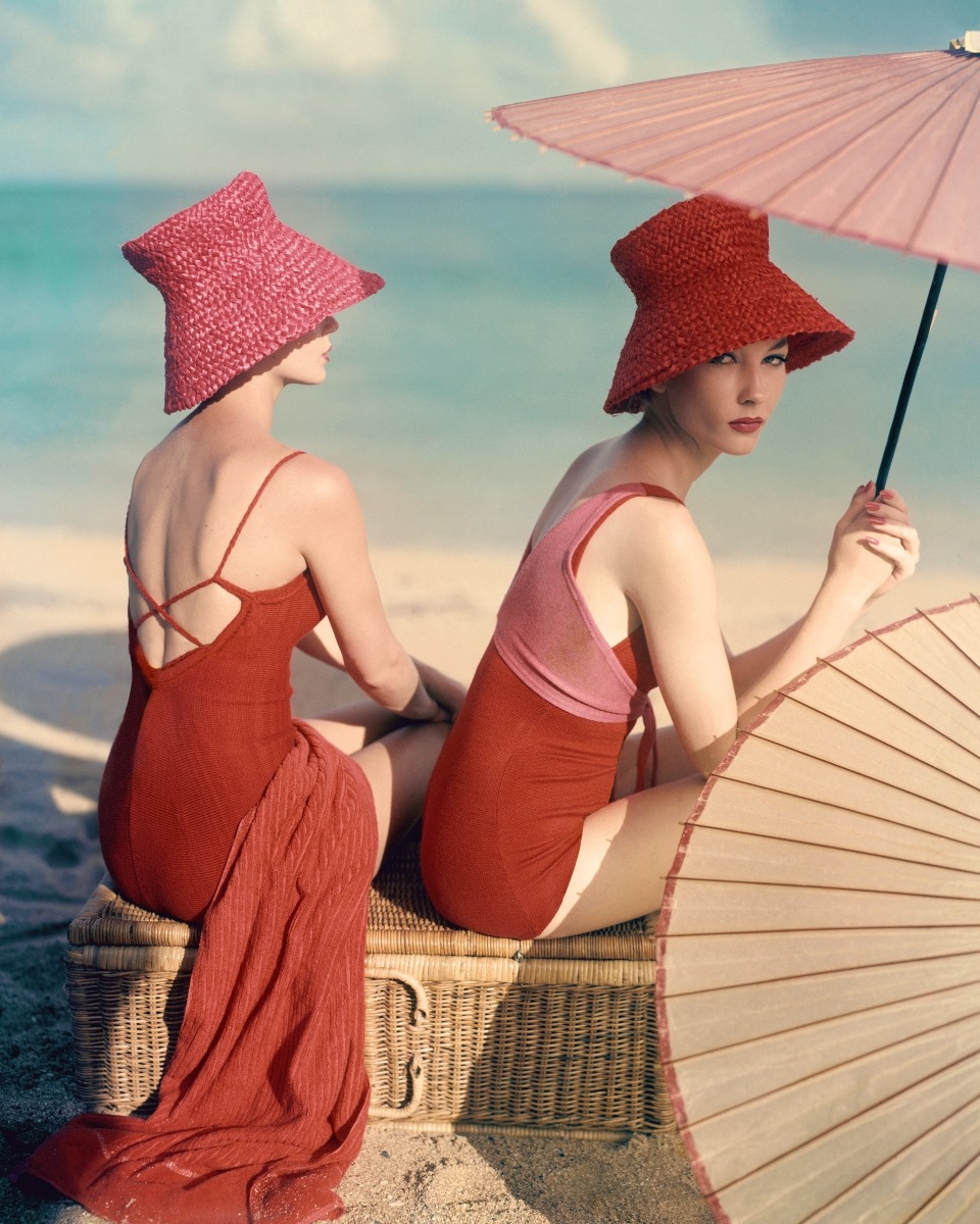 Two models seated on a large wicker picnic basket, with silk and bamboo umbrellas, wearing swimsuits and straw hats. Left: Red wool and nylon swimsuit with thin criss-crossed straps over a low-scooped back, by Margaret Pennington, a pink cylinder-topped conical straw hat by Emme, and a ribbed pink towel by Wamsutta. Right: Red swimsuit wth a layer of pink strapping across the top, tied below the chest, of elasticized red wool, by Gernreich-Westwood, with a red cylinder-topped conical straw hat by Emme.