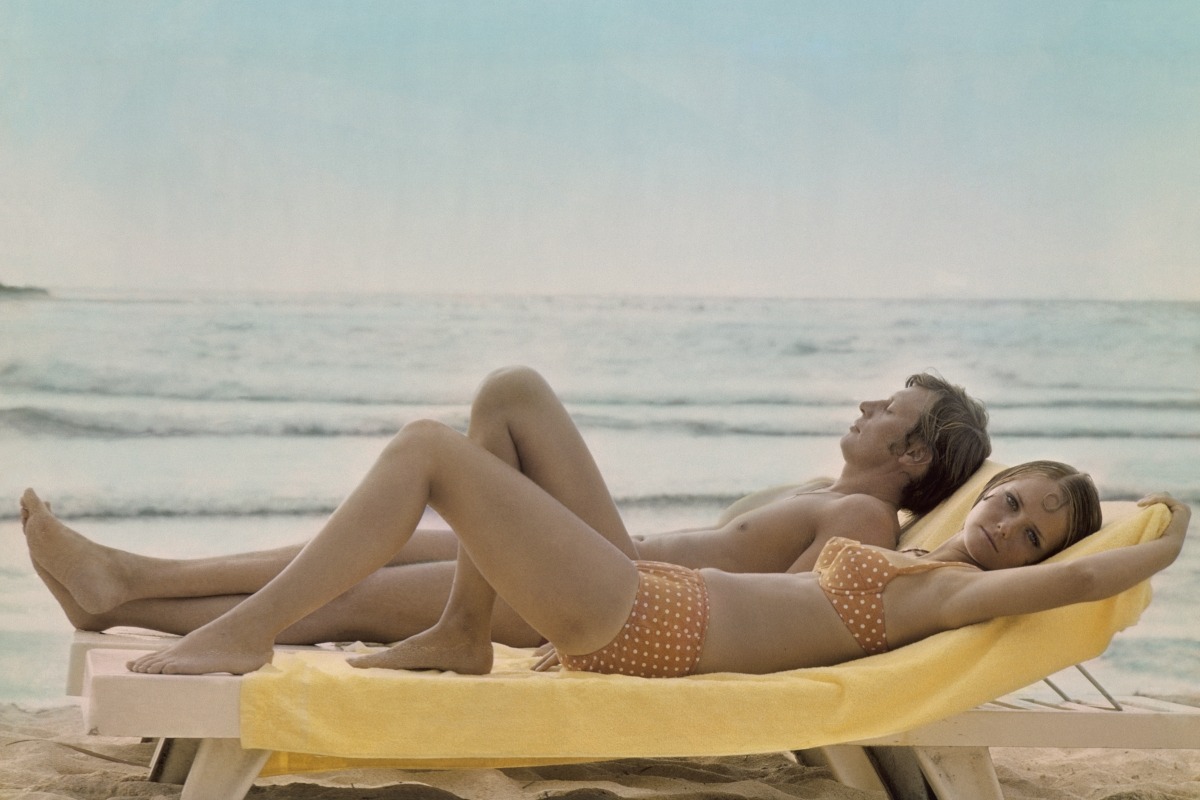 Model Cheryl Tiegs at the beach in an orange bikini with white polka dots by Villager, with a man reclining on chaise 