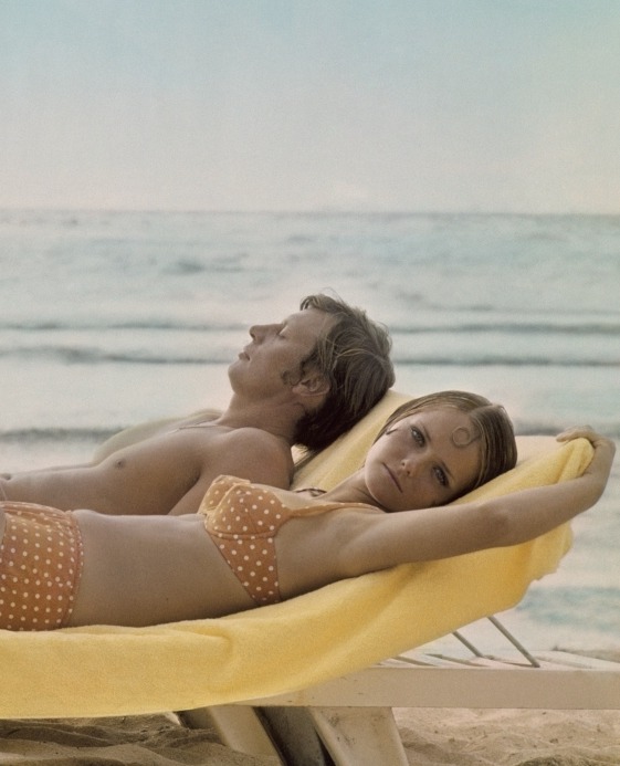 Model Cheryl Tiegs at the beach in an orange bikini with white polka dots by Villager, with a man reclining on chaise 