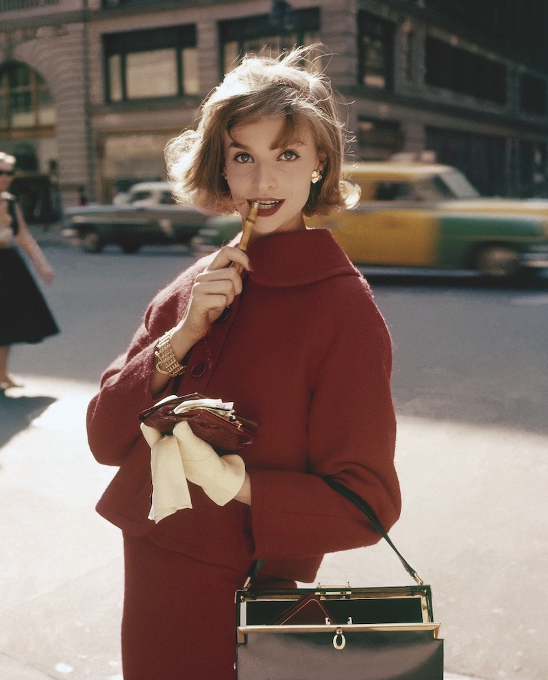 Shopping for Christmas: Model, holding pencil to mouth while standing on New York street, in red suit, white gloves and open handbag 