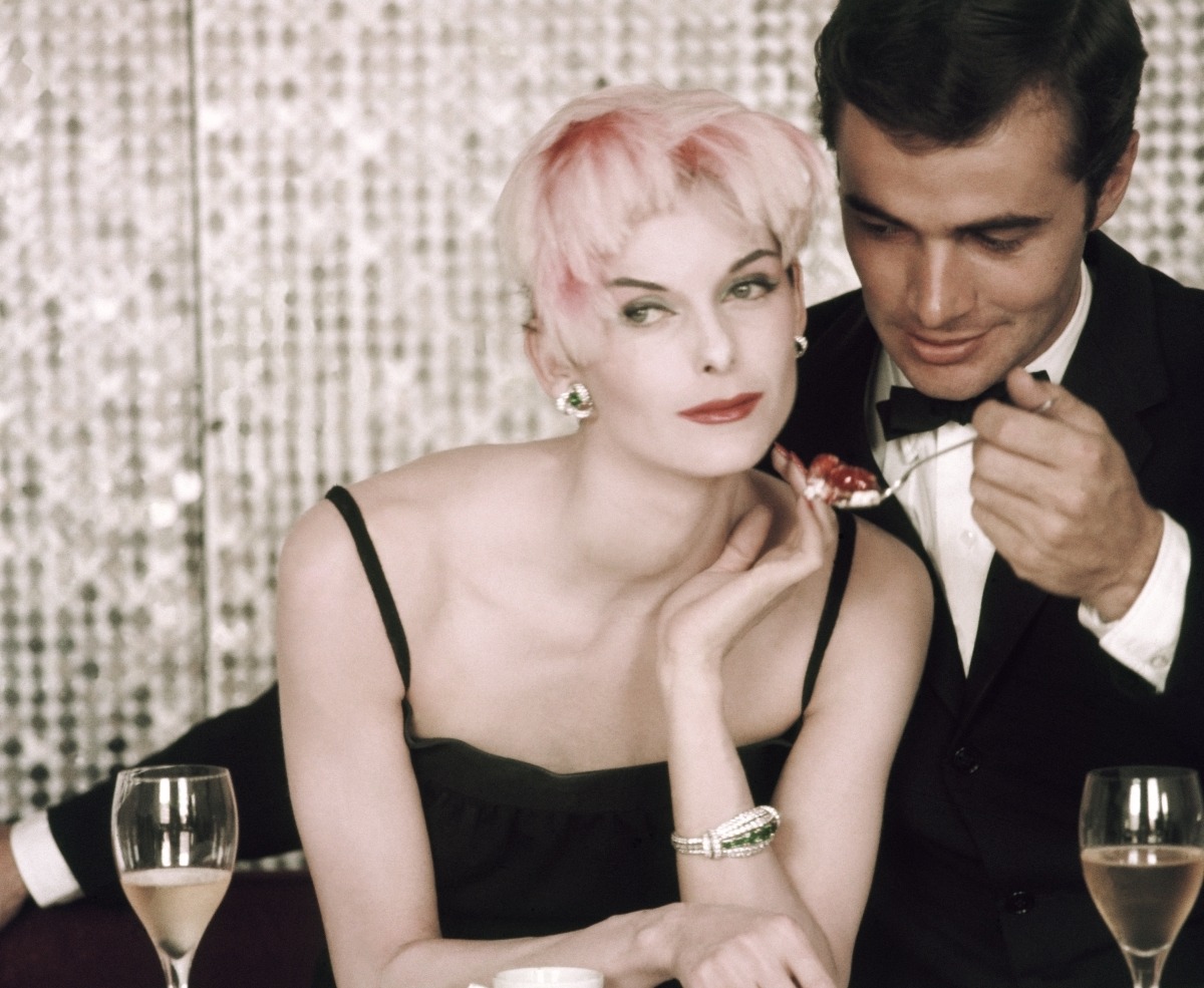 Model Anne St. Marie poses with a male model at a dinner party table, a cup of espresso in front of her and two glasses of wine on either side as the male model offers her a spoonful of food. St. Marie is wearing a black dress, a flamingo pink feathered cap, and earrings of diamonds and emeralds by Cartier. Makeup using Elizabeth Arden products from the "Oriental Pink" line. *** Local Caption *** Anne St. Marie;