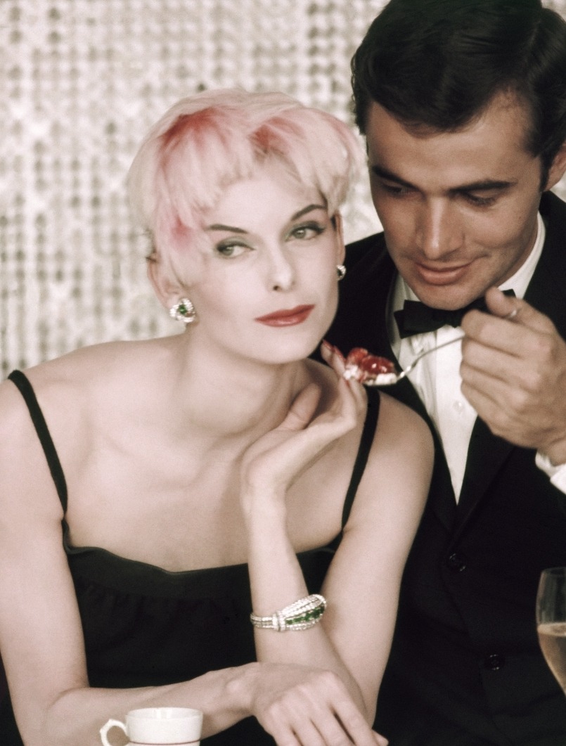 Model Anne St. Marie poses with a male model at a dinner party table, a cup of espresso in front of her and two glasses of wine on either side as the male model offers her a spoonful of food. St. Marie is wearing a black dress, a flamingo pink feathered cap, and earrings of diamonds and emeralds by Cartier. Makeup using Elizabeth Arden products from the "Oriental Pink" line. *** Local Caption *** Anne St. Marie;