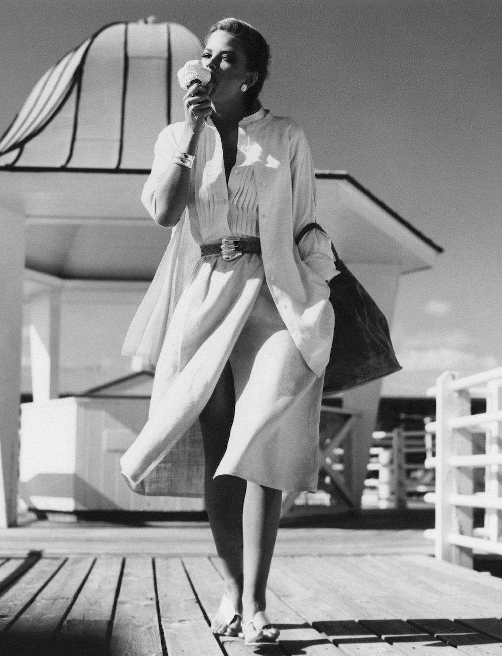 Model, Lisa Taylor, walking at the Westhampton Bath and Tennis Club, in Long Island, New York, eating an ice cream cone, and wearing a long linen collar-band shirt-jacket, a white linen blouse and wrap skirt by Calvin Klein, with a large leather bag on left arm. (Photo by Albert Watson/Conde Nast via Getty Images) Vogue 1976