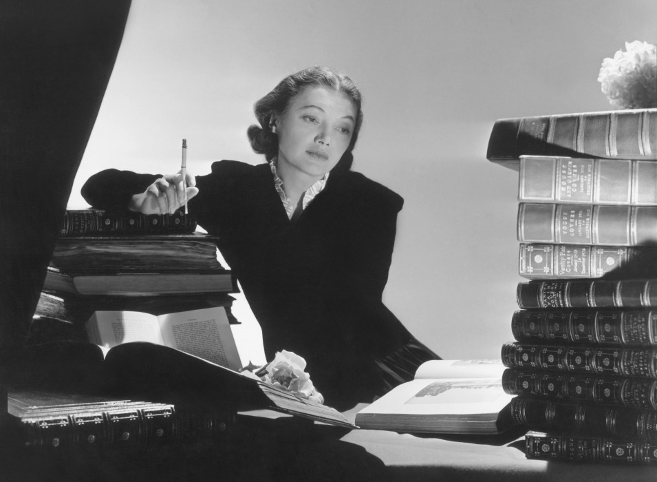 Actress Katharine Cornell wearing a black jacket, white ruffles showing at the neckline, surrounded by books *** Local Caption *** Katharine Cornell; Vogue February 01, 1938 Portrait