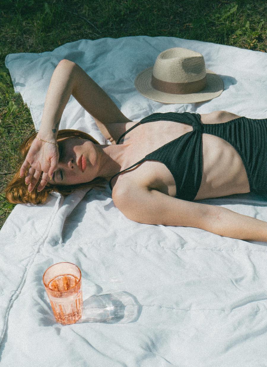 žena leži na travi i sunča se u crnoj haljini, a pored nje je čaša vode za hidrataciju