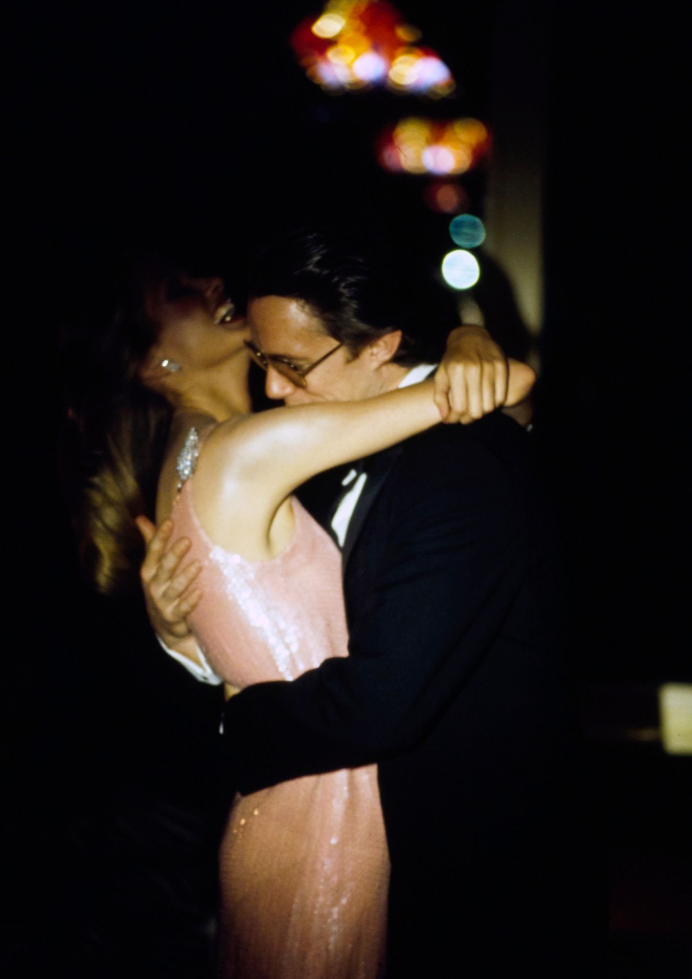 Model embracing a male model with multi-colored lights shining behind them. She is wearing a long evening dress of light pink sequins on silk chiffon by Halston Ltd