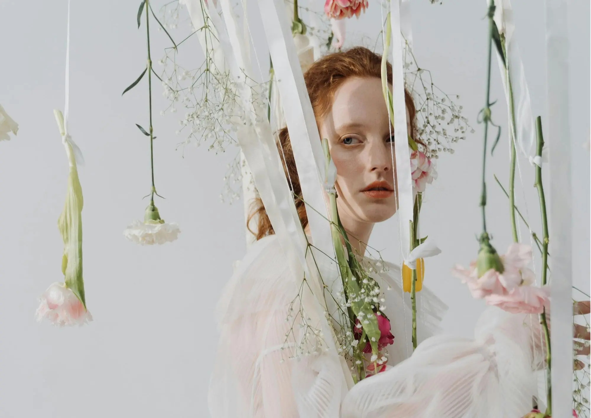 žena s crvenom kosom u bijeloj bluzi okružena cvijećem koje visi sa svilenih vrpca