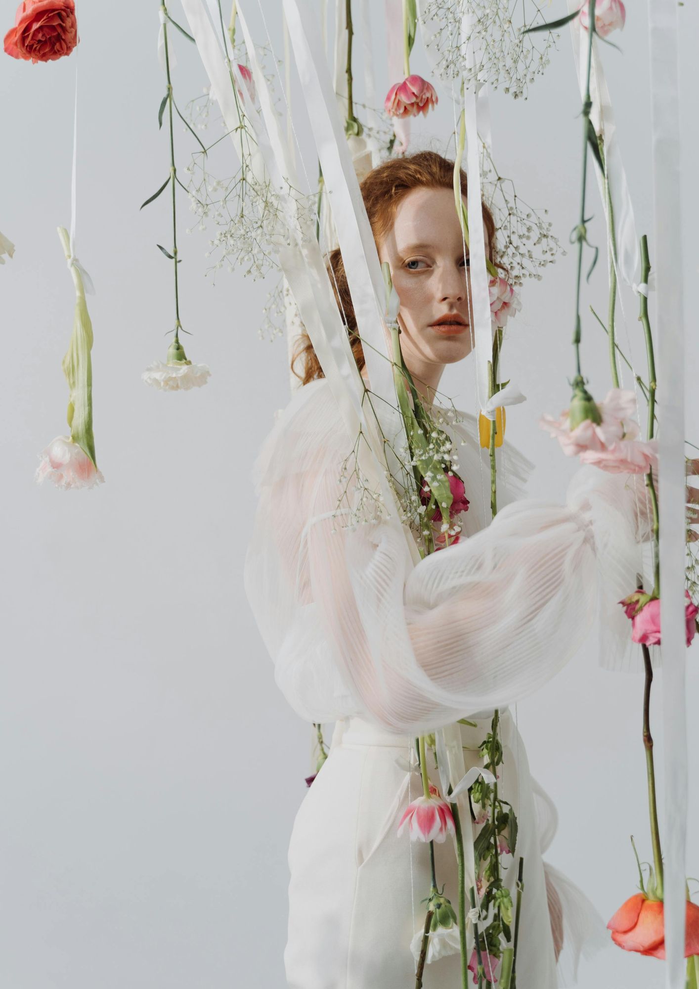 žena s crvenom kosom u bijeloj bluzi okružena cvijećem koje visi sa svilenih vrpca