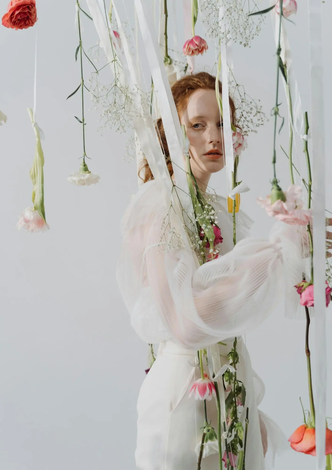 žena s crvenom kosom u bijeloj bluzi okružena cvijećem koje visi sa svilenih vrpca